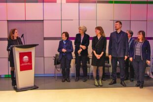 One woman speaks from the podium with five people standing to her left.